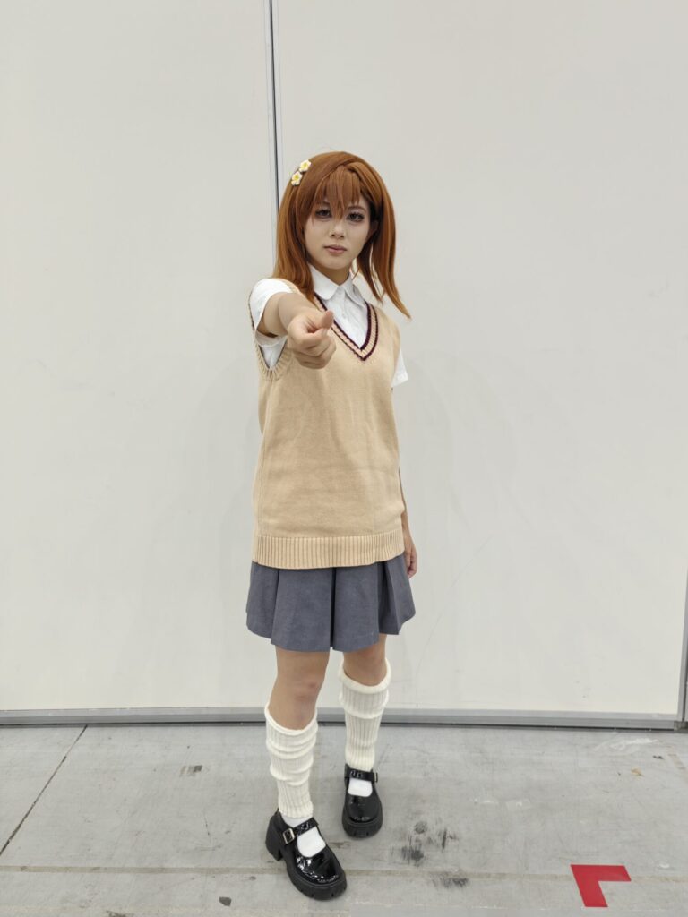 Cosplayer caracterizada como Mikoto Misaka (Railgun), com uniforme escolar e gesto confiante.